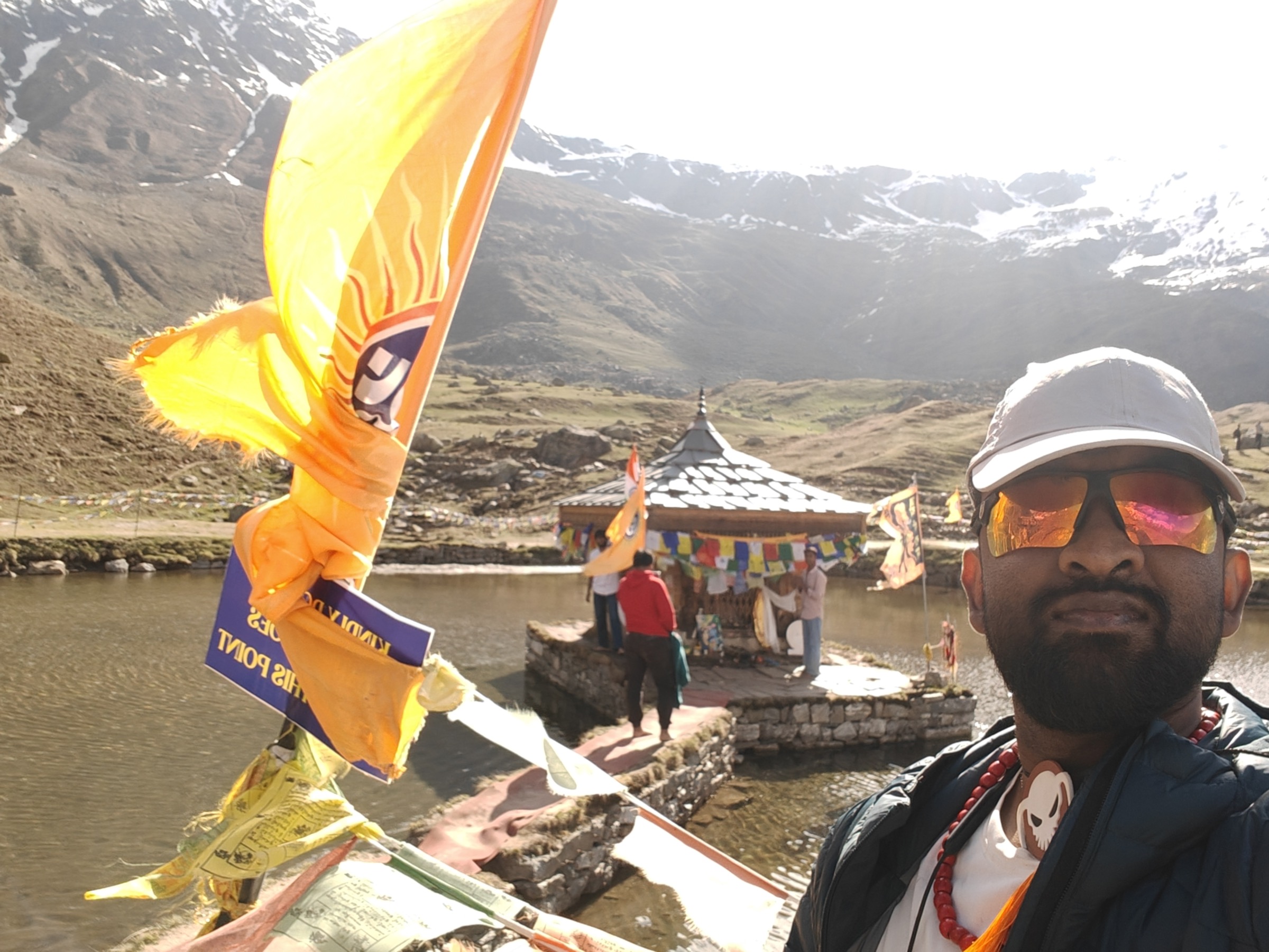 Karthikeyan at a Himalayan lake temple with prayer flags and snow-capped peaks, on a trek between race blocks